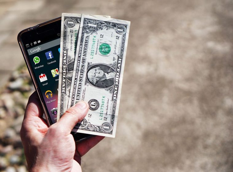 Mão segurando um smartphone com aplicativos abertos ao lado de notas de dólar, em ambiente externo.