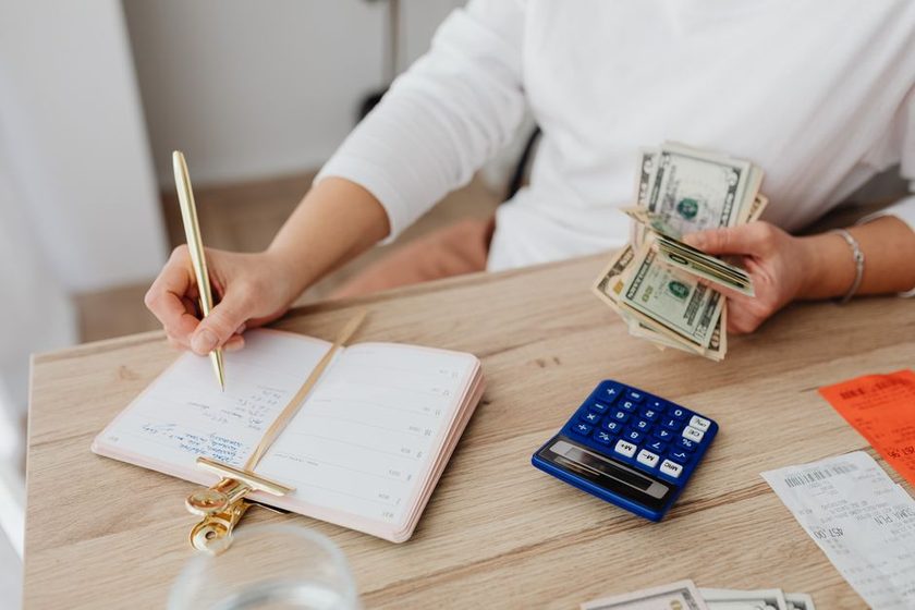 Mulher anota compromissos numa agenda de papel enquanto segura notas de dinheiro ao lado de uma calculadora, sobre uma mesa de madeira.